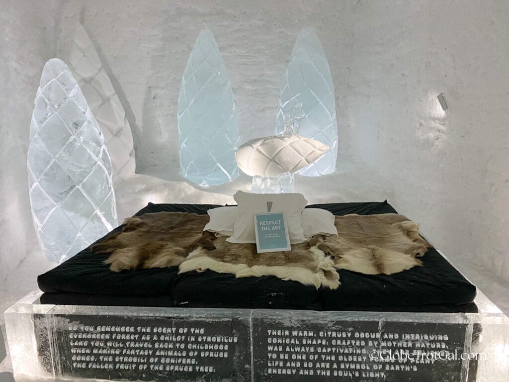 Large pinecones carved of ice surrounding a bed with an ice frame and a mattress covered with reindeer hides and pillows with a sign that says "respect the art".