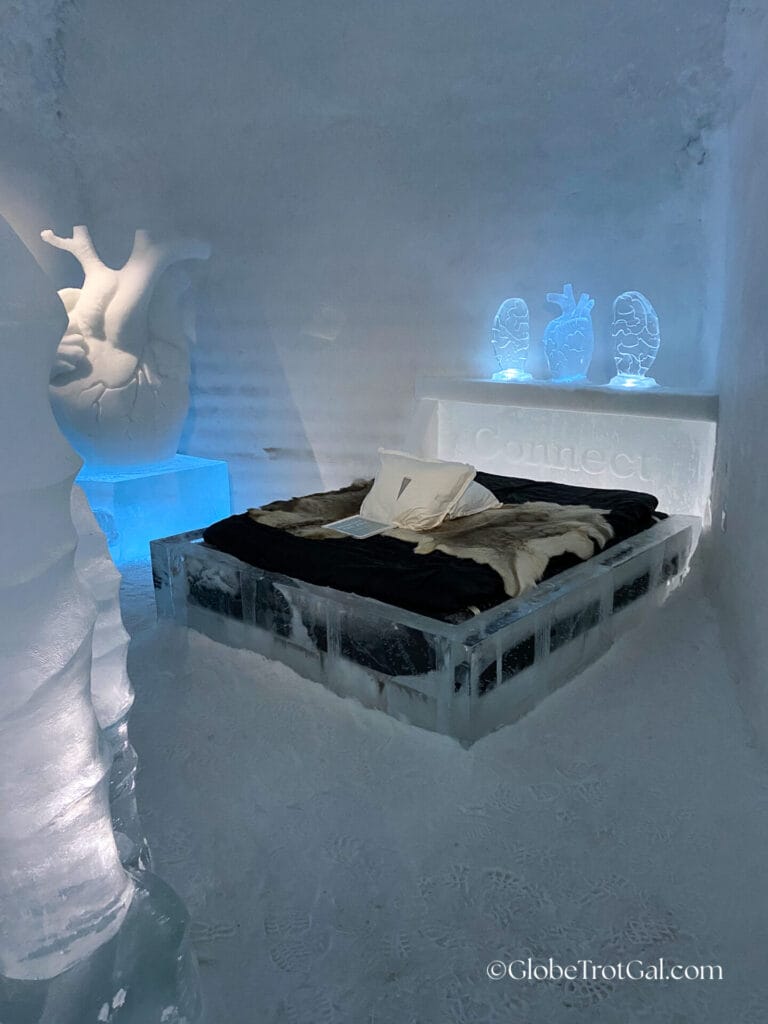 Ice room with ice sculptures of a brain and heart and a bed frame made of ice that says "Connect" on it, with a mattress covered in reindeer hides.