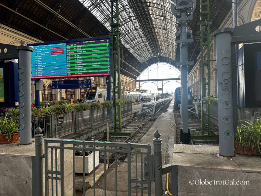 Interior of main train hall in Budapest Keleti station with arched glass roof, train tracks, and digital board showing train schedules.