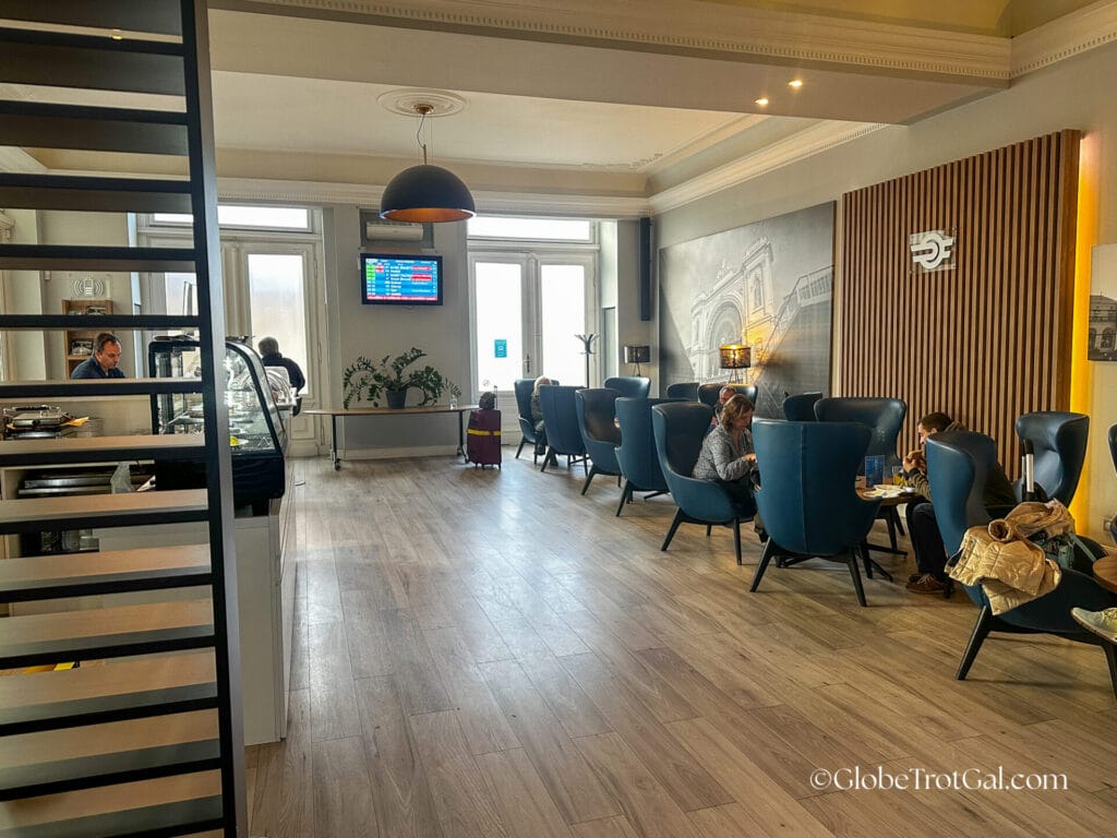 Interior of Budapest Keleti Station First Class Lounge with a wooden floor, blue leather chairs in sets of four and a TV screen on the back wall showing train schedules.