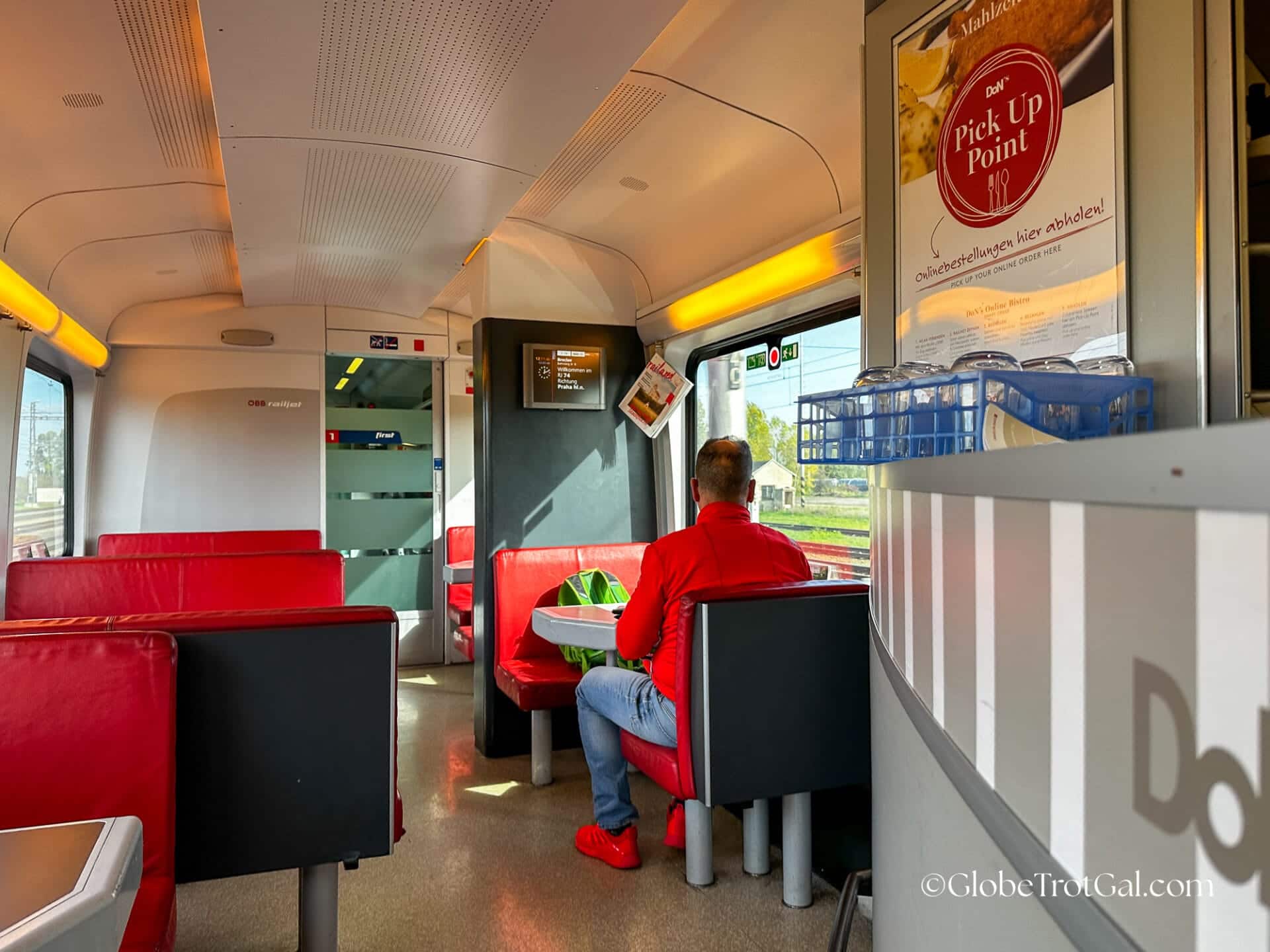 View down aisle of dining car on OBB Railjet train with red booths on either side of the aisle and man in a red shirt sitting on the righthand side.