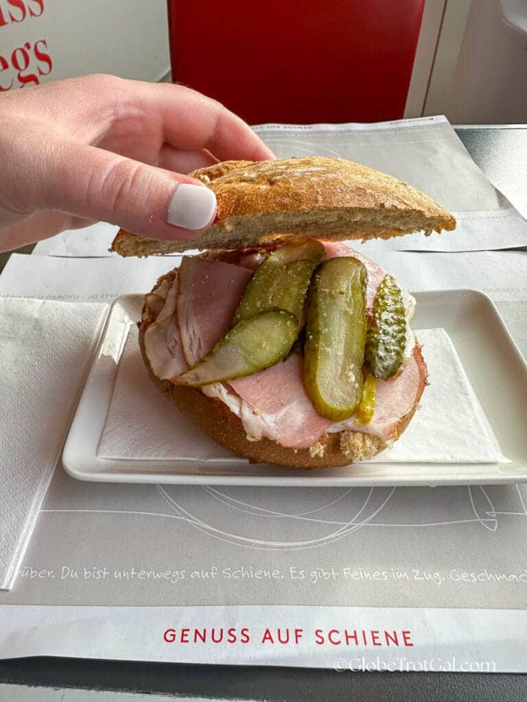 A hand picking up the top bun of a sandwich to show ham and pickles.