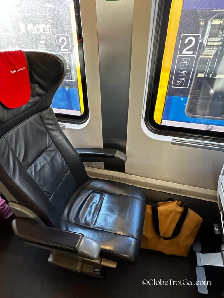 Black leather first class train seat with a red headrest and a yellow tote bag sitting on the floor in front of it on an OBB Railjet train.