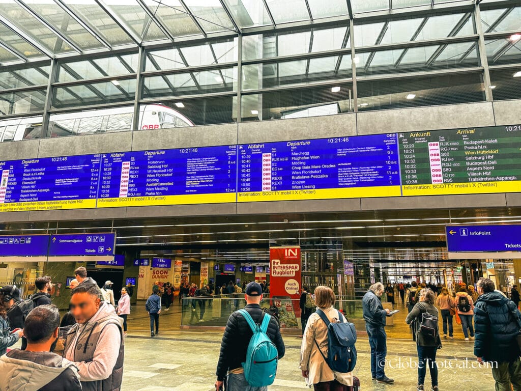Main hall at Vienna HBF (central train station) with passengers walking around and large blue digital train schedule display overhead.