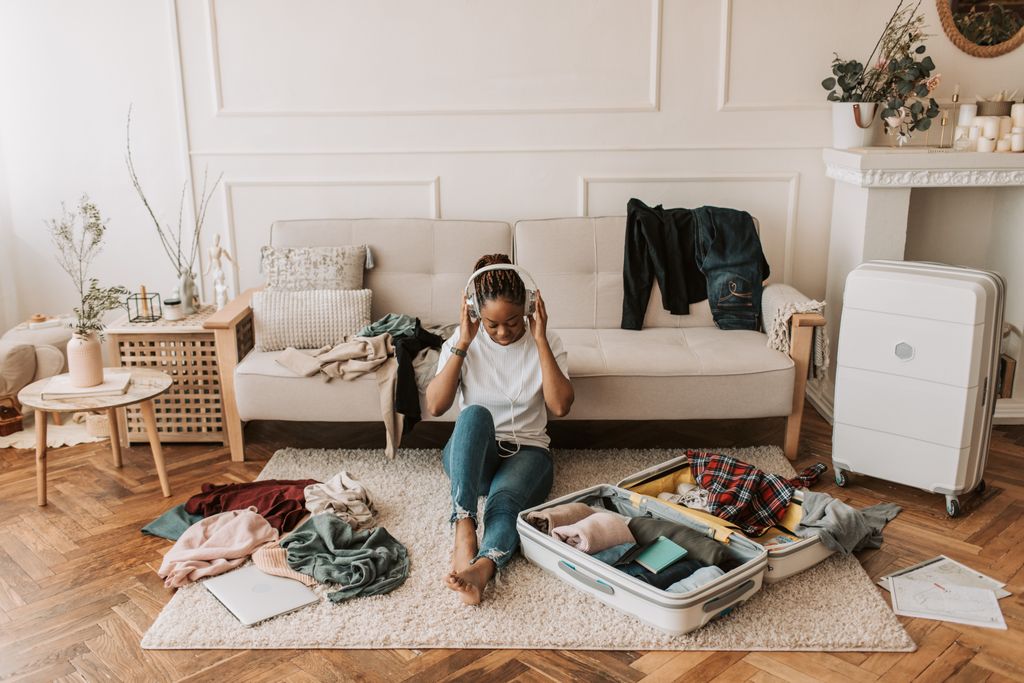 A woman packs a suitcase in her living room while listening to music, surrounded by clothes and a cozy atmosphere.